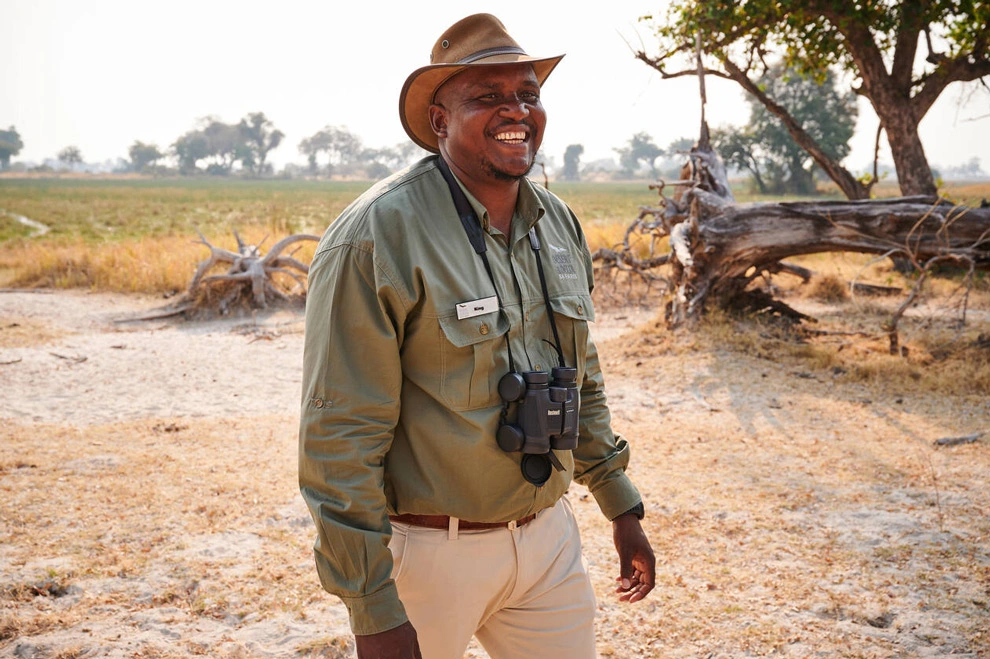 Guide de safari souriant, équipé de jumelles et d’un chapeau, debout dans un paysage de savane avec arbres et herbes sèches en arrière-plan.