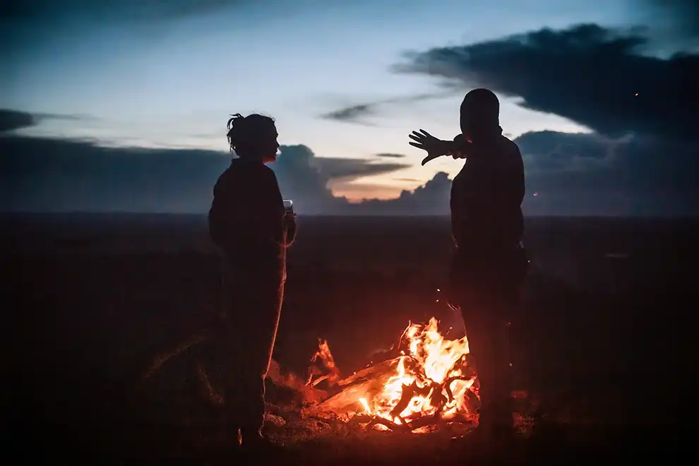 Un voyageur et un guide échangent près d’un feu de camp au coucher du soleil, symbolisant la rencontre et la convivialité au cœur d’un voyage en Afrique.