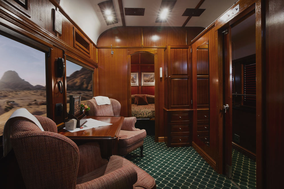 Un compartiment de train élégant en bois avec fauteuils, bureau et cabine de couchage, offrant une vue sur un paysage désertique.
