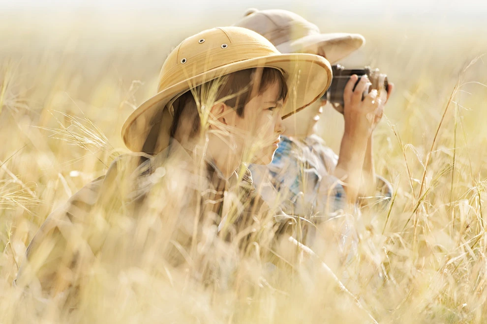Enfants avec des chapeaux de safari observant la nature dans les hautes herbes et des jumelles.