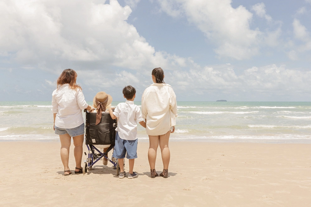 Famille avec une personne en fauteuil roulant face à l’océan sur une plage de sable.