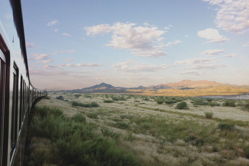 Un train longe une vaste plaine herbeuse dans le Kalahari avec des montagnes à l’horizon sous un ciel parsemé de nuages.