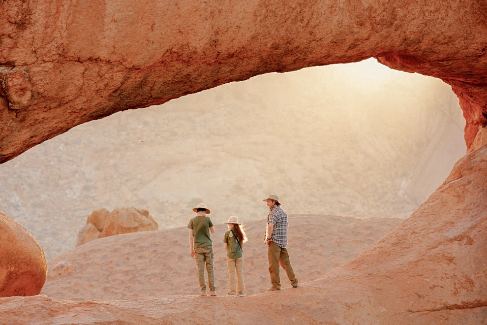 Famille explorant le paysage désertique de Namibie sous une arche rocheuse naturelle de Spitzkoppe au coucher du soleil.