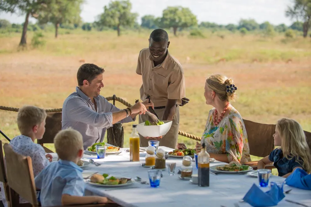 Une famille déjeune en plein air dans la savane en Zambie pendant qu’un serveur leur apporte un plat.