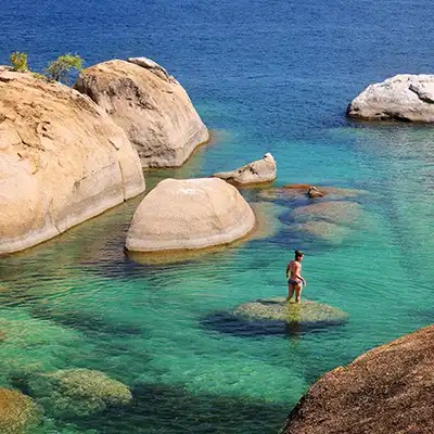 Voyageuse marchant entre les rochers dorés dans les eaux turquoise du lac Malawi, un paysage paisible et lumineux au cœur de l’Afrique australe.