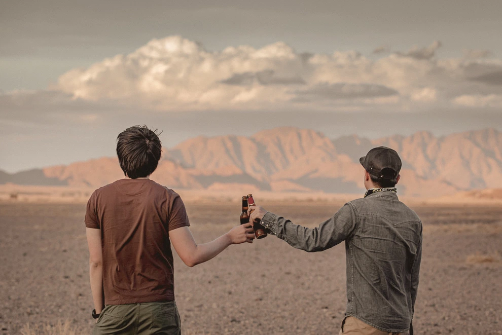 Deux voyageurs trinquant avec des bouteilles face à un paysage désertique spectaculaire du Damaraland en Namibie avec des montagnes au loin.