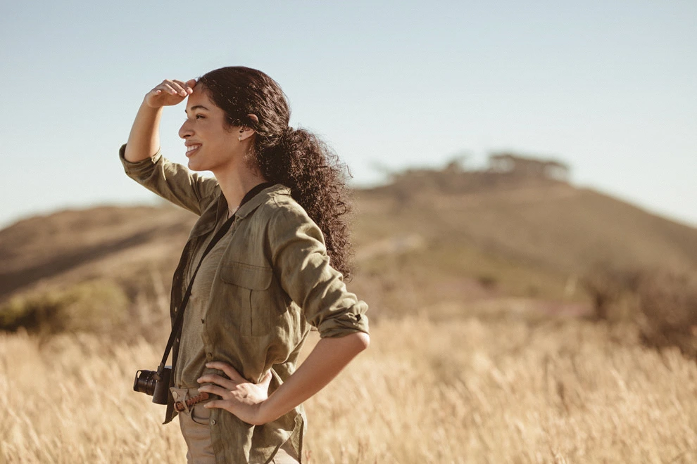Femme en tenue de safari scrutant l’horizon dans une savane dorée avec un appareil photo autour du cou.