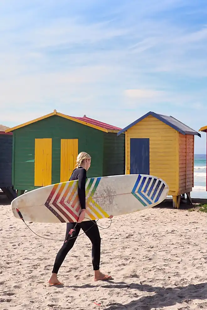 Surfeuse marchant sur la plage de Muizenberg en Afrique du Sud, devant les cabanes colorées emblématiques.