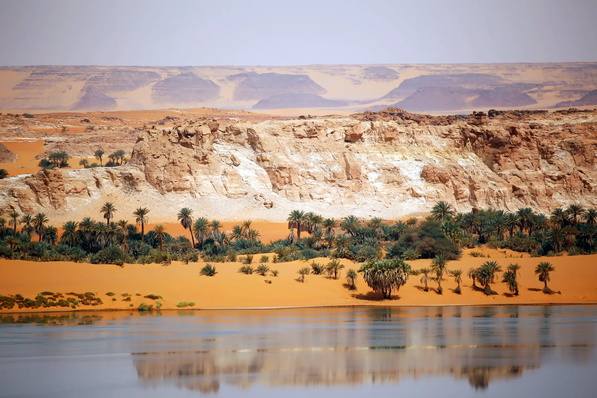 Paysage du Tchad : dunes ocre, palmeraie et lac du Sahara reflétant les falaises dorées.