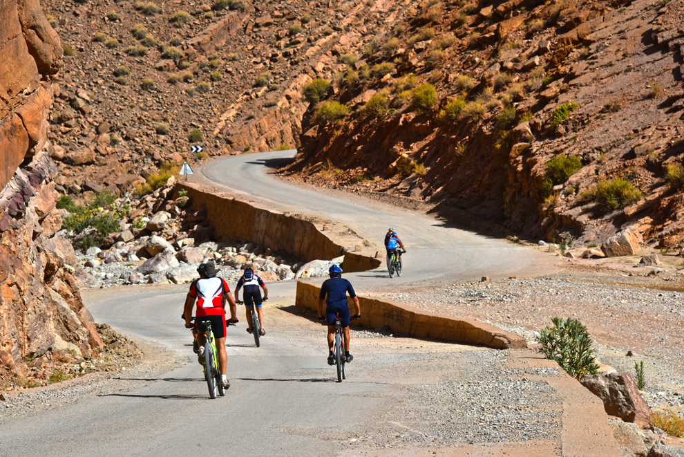 Quatre cyclistes progressent sur une route sinueuse au Maroc à travers un paysage rocheux et aride.
