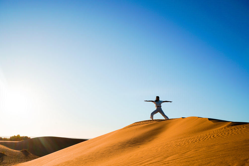 Une personne en tenue claire pratique une posture de yoga au sommet d’une dune de sable.