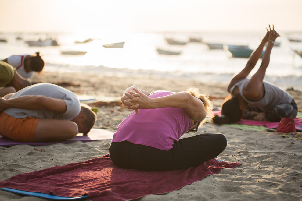 Un groupe de personnes pratique le yoga sur une plage au coucher du soleil, avec la mer et des bateaux en arrière-plan.