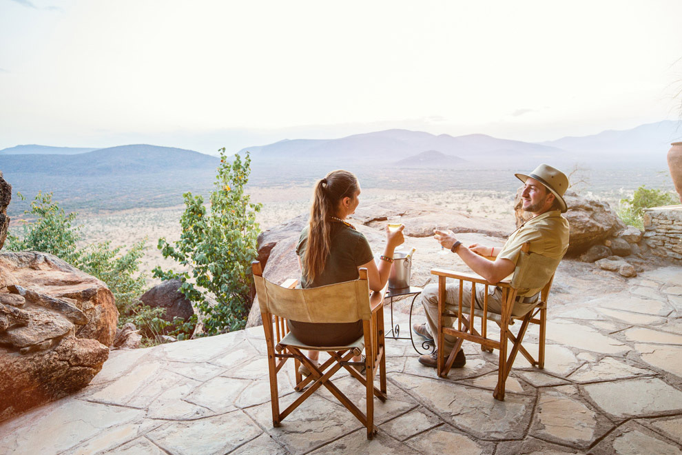 Couple profitant d’un apéritif face à une vaste vue sur la savane et les montagnes depuis une terrasse en hauteur.