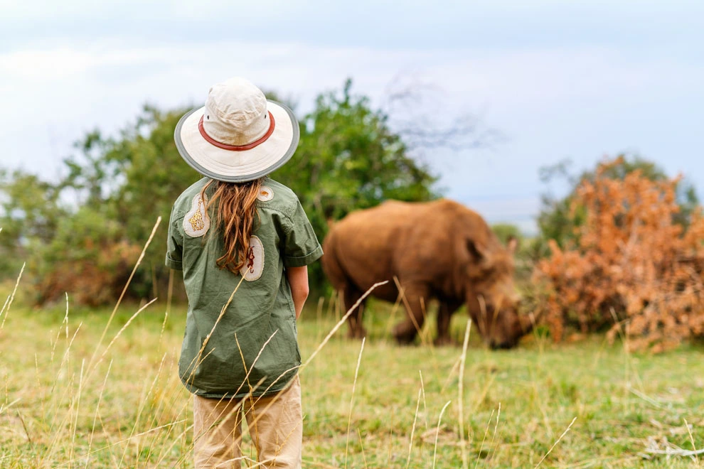 Enfant en tenue de safari observant un rhinocéros qui broute dans la savane.