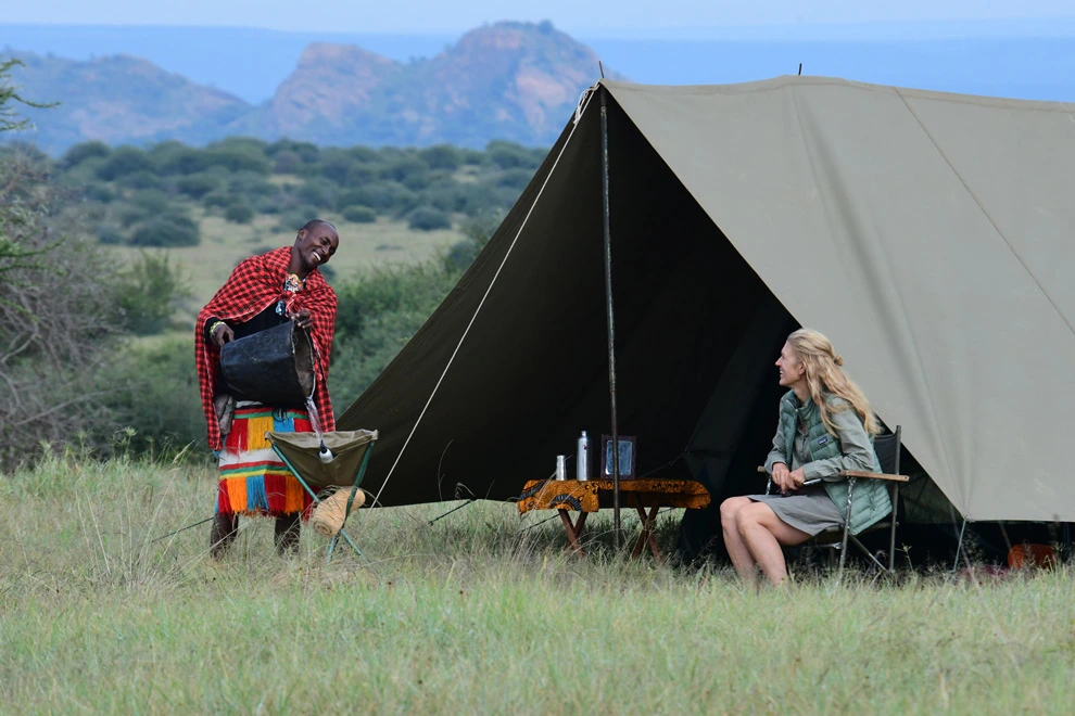 Campement de toile en savane, avec un guide Samburu versant de l’eau à une jeune femme.