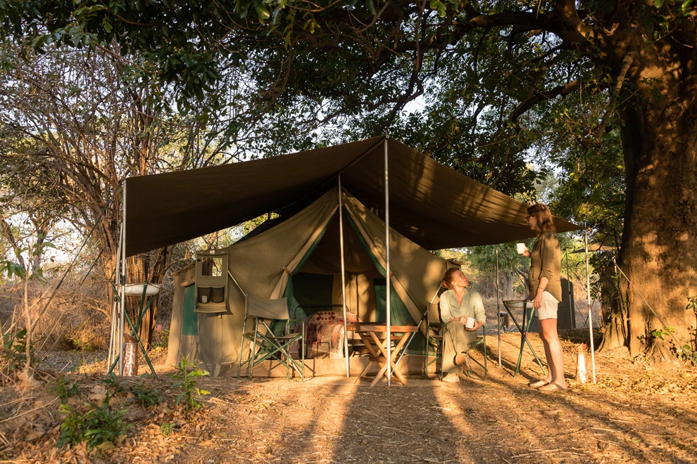 Tente de safari installée à l’ombre d’arbres avec deux voyageurs.
