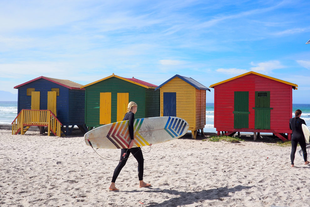 Surfers devant des cabanes de plage colorées sur une plage en Afrique du Sud.