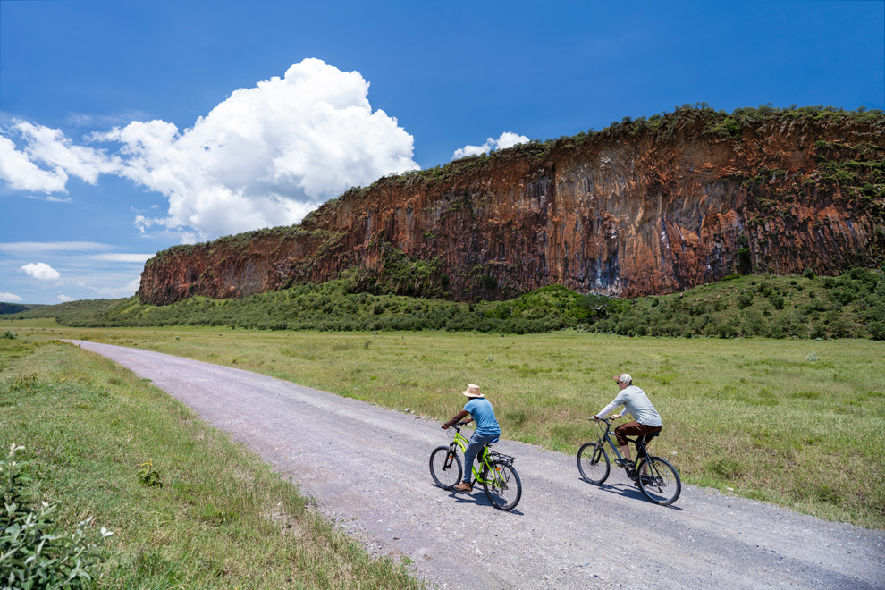 Deux cyclistes roulent sur une piste au pied d’une falaise rocheuse dans la savane.